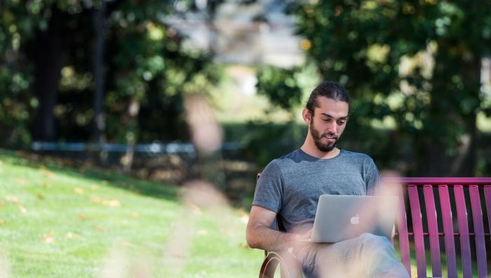 A student studies on his laptop outdoors on a bench.