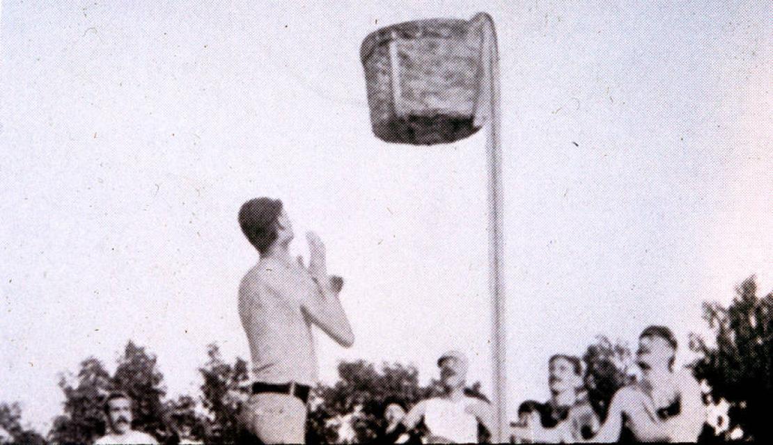 An early outdoor basketball game using a peach basket is played at Springfield College.