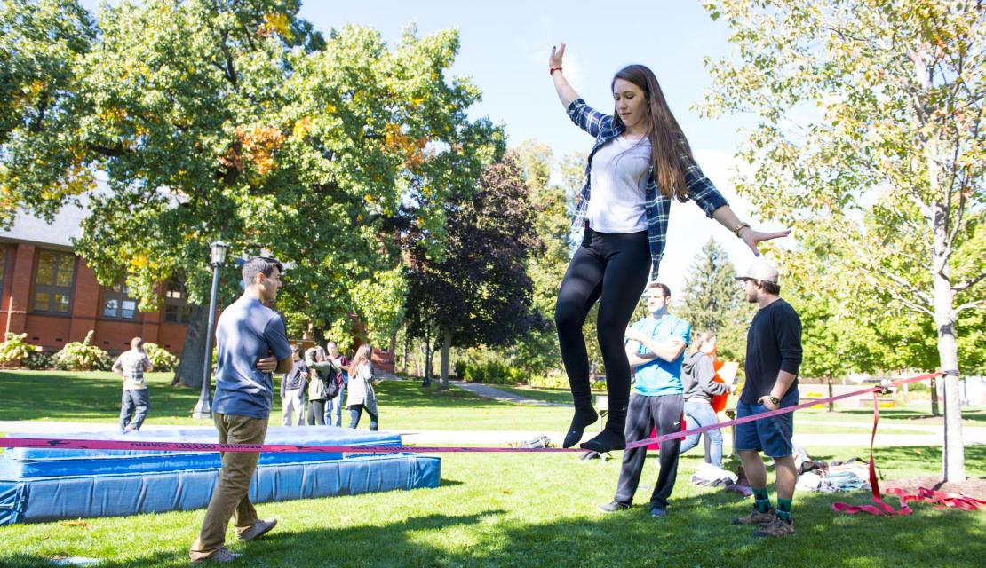 A female student walks across a tight rope on the campus green. 