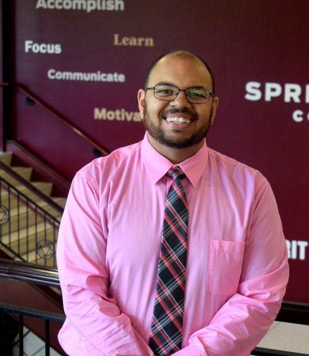 A photo of Robert Gordon, smiling, standing in front of a wall with the Springfield College logo on it