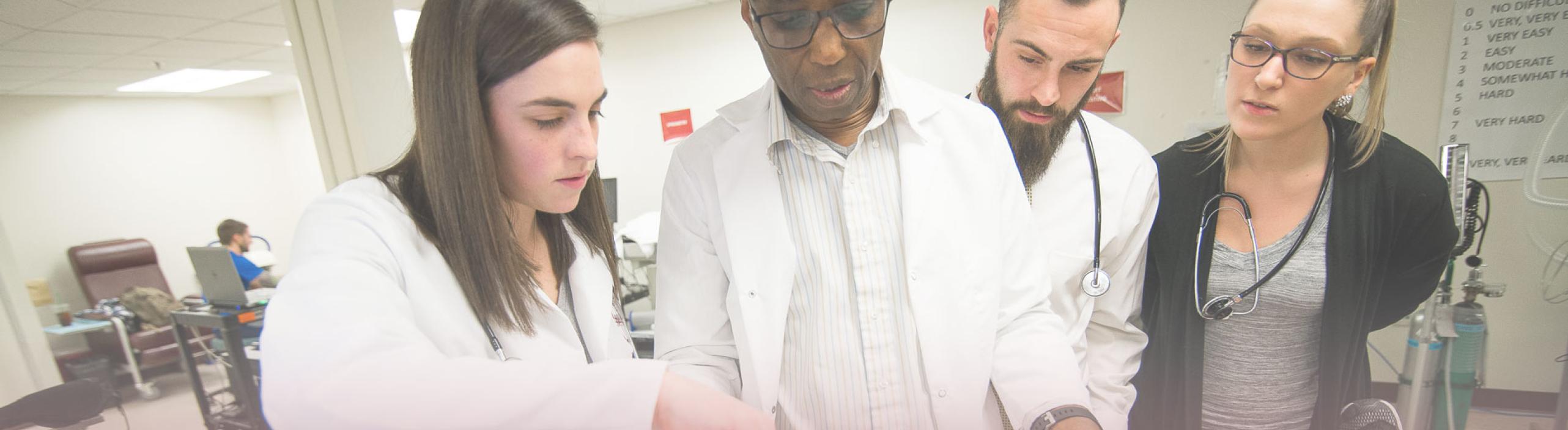 Three graduate students look on as a professor leads a class in a lab