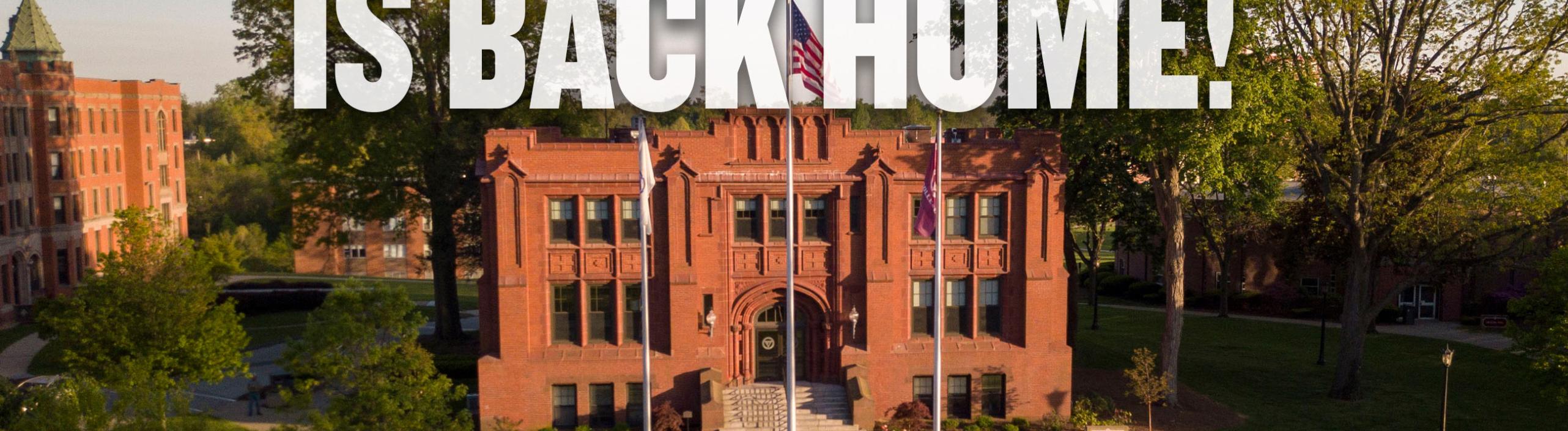Img: A drone shot of the flag poles in front of Marsh Memorial on the Springfield College Campus, with a text overlay in white saying "Homecoming Weekend is Back Home!"