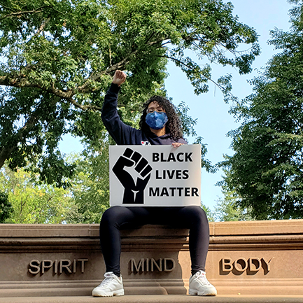 student sitting on spirit mind body wall at Springfield College with raised fist and BLM sign