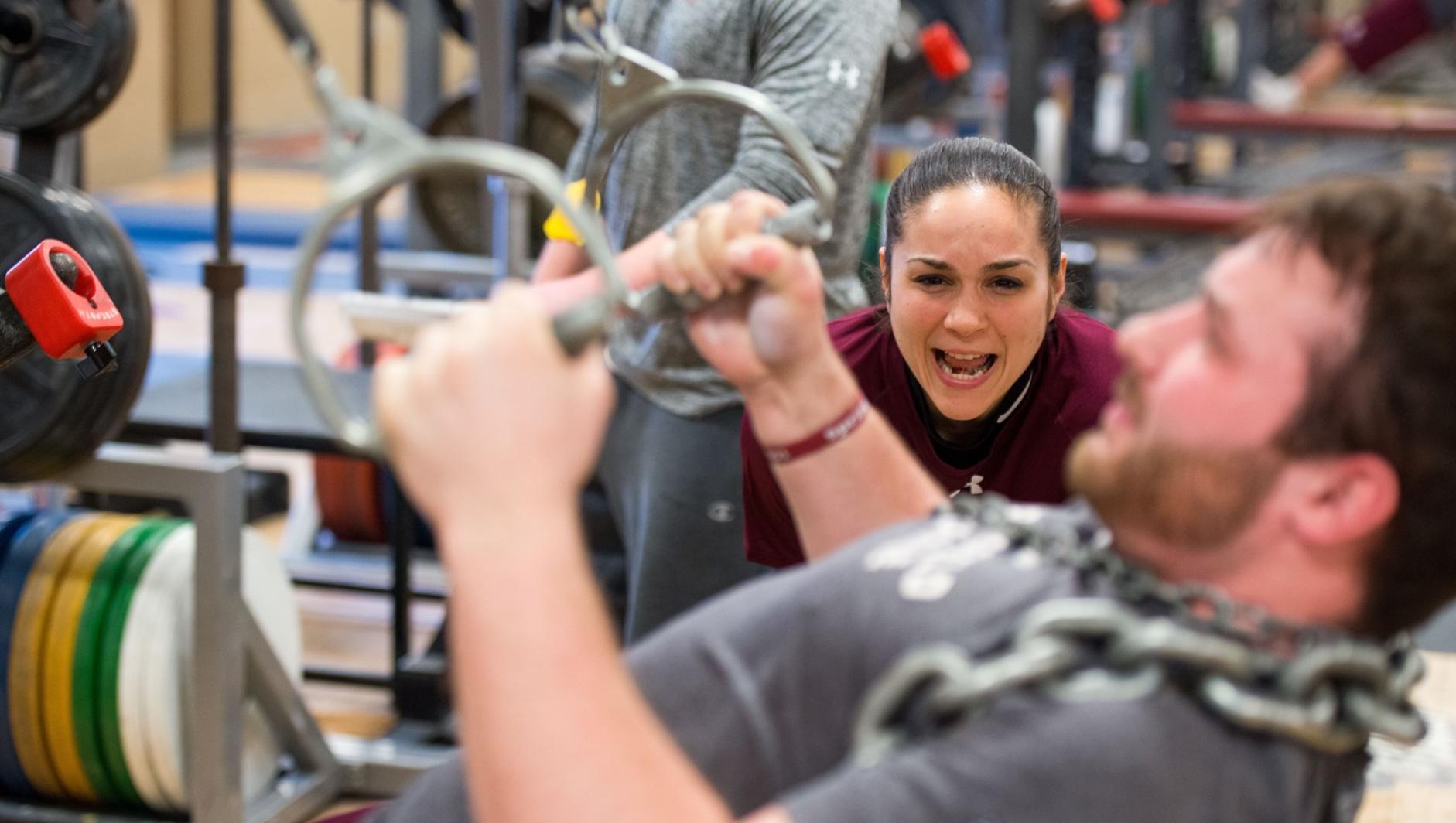 A student strength and conditioning coach yells words of encouragement as a student works out his arms.