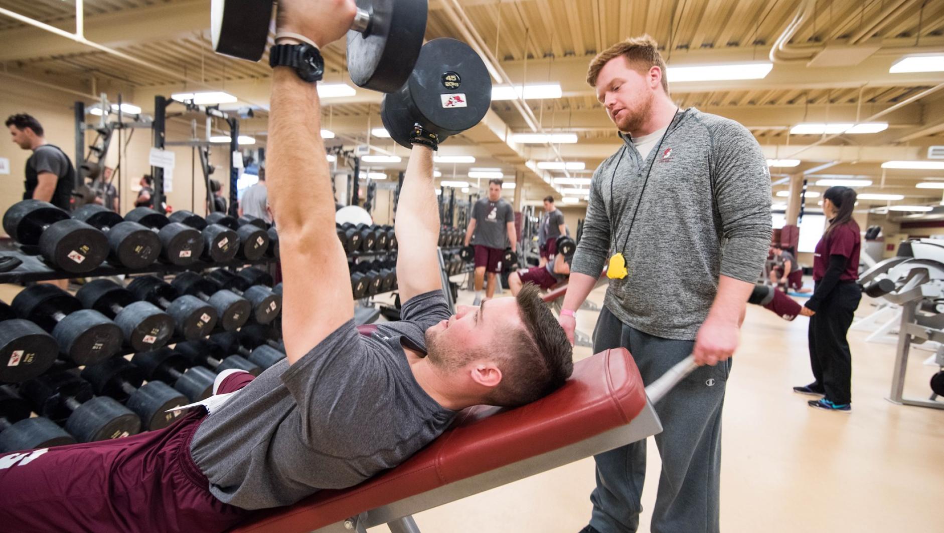 A student strength and conditioning coach looks on as his client trains with dumbbells.
