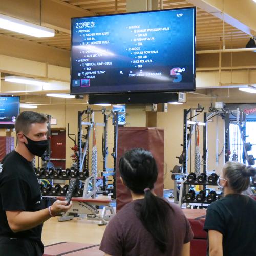 a male student. wearing a mask, is explaining how to use the new digital screens in the strength and conditioning room to two female student-athletes. The new screen is visible in the background.
