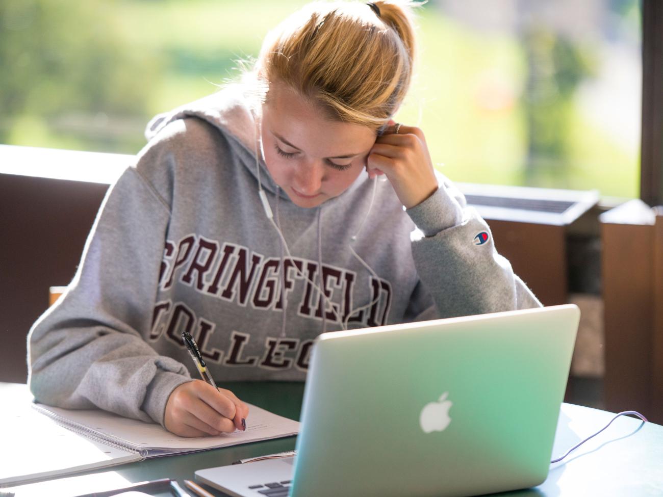 A student writes in a notebook at a desk with a laptop open in front of her.
