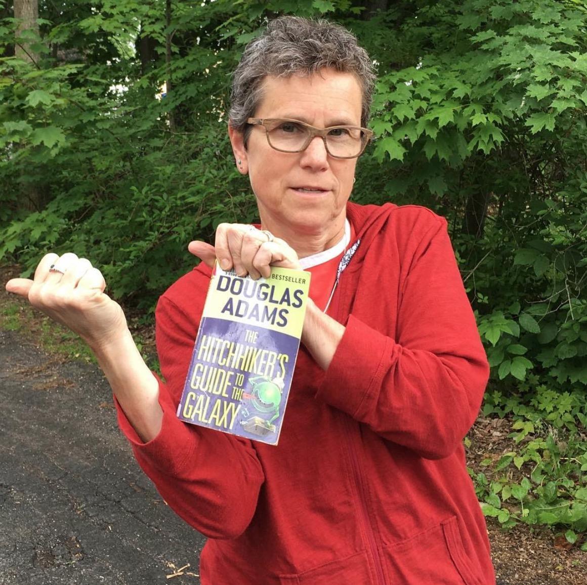 Julia Chevan jokingly hitchhikes while holding a copy of Hitchhikers Guide to the Galaxy
