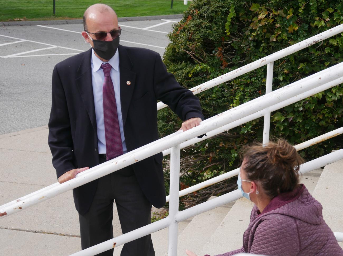 Criag Poisson, wearing a mask, is standing on the stairs of Blake Arena talking with Women's Lacrosse Head Coach Kristen Mullady, who is sitting on the steps