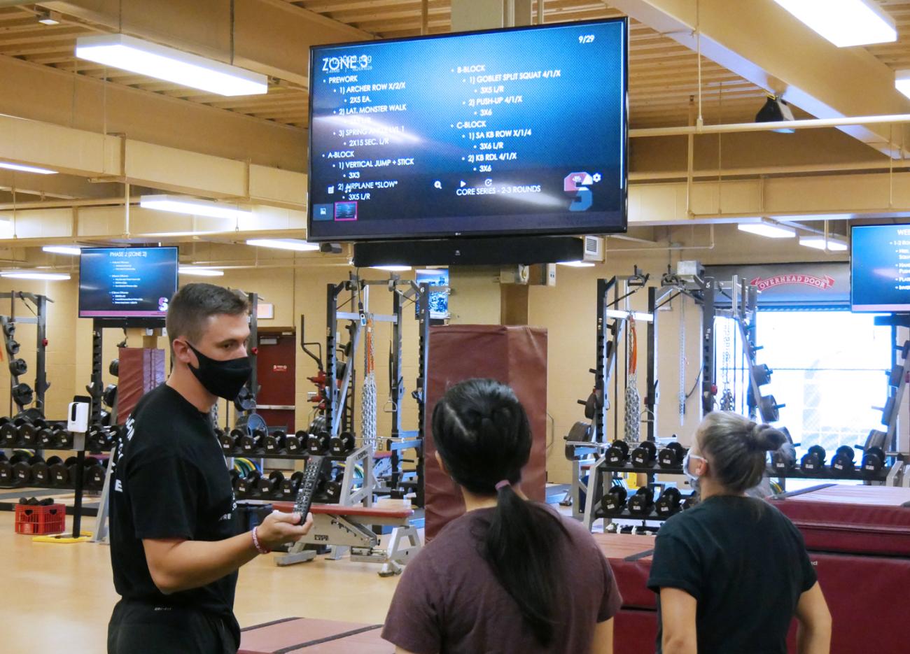 a male student-athlete, wearing a mask, is showing two female student-athletes how to use the new digital screens in the strength and conditoning room