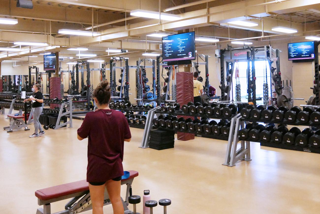 A view of the weights section of the strength and conditioning room, with weight machines and new digital screens, a female student-athlete is standing to the left at a weight bench