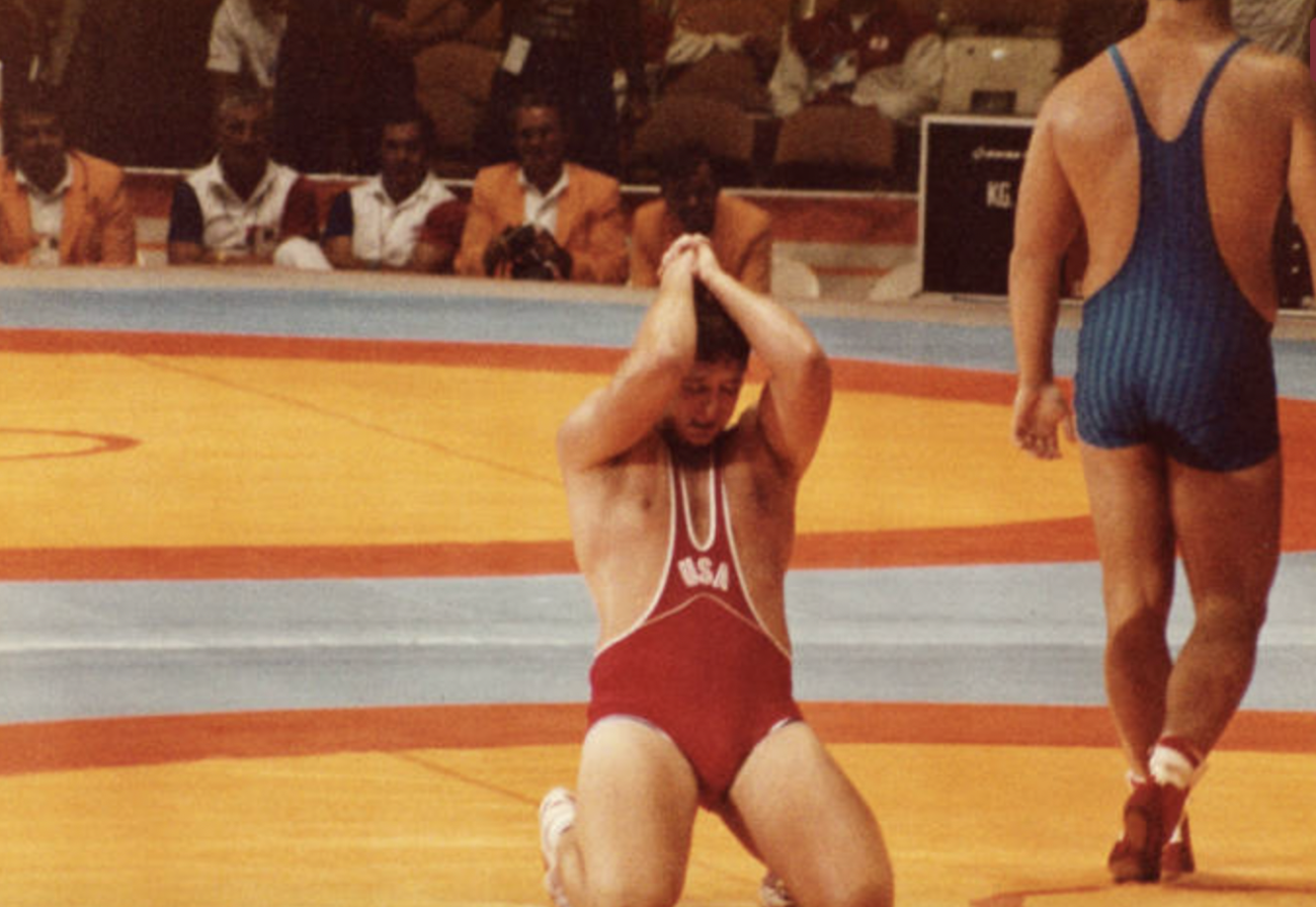 Blatnick kneeling on the floor during a wrestling match as his opponent is walking away. He is wearing a USA Olympic wrestling uniform. The picture may have been taken during the 1984 Olympics, where he won a Gold medal.