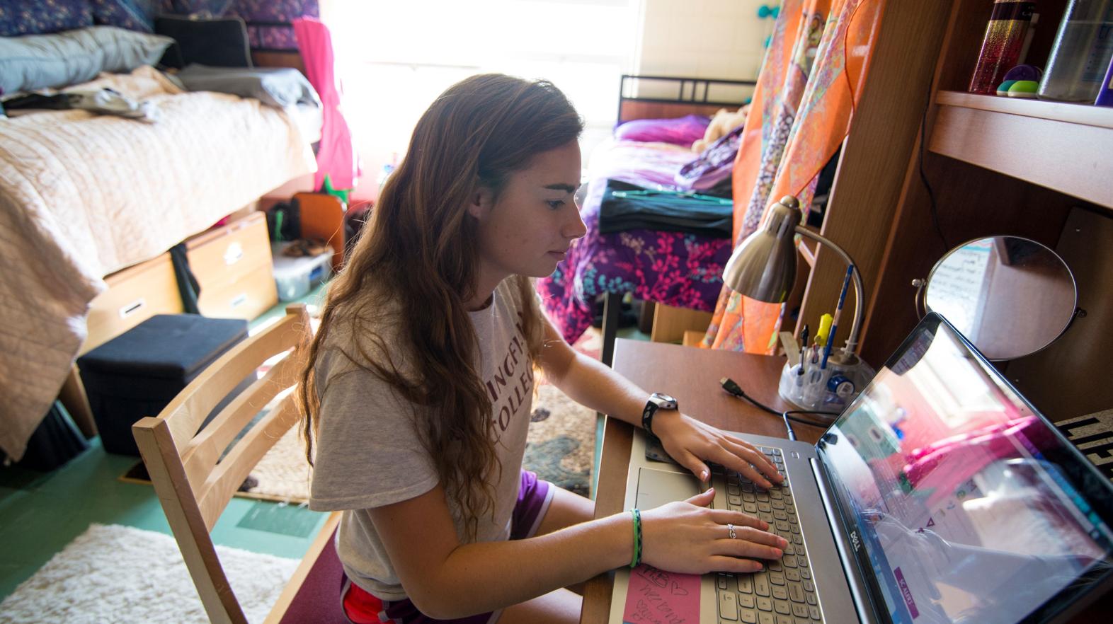 A student sits at her desk in her residence hall working on her laptop.