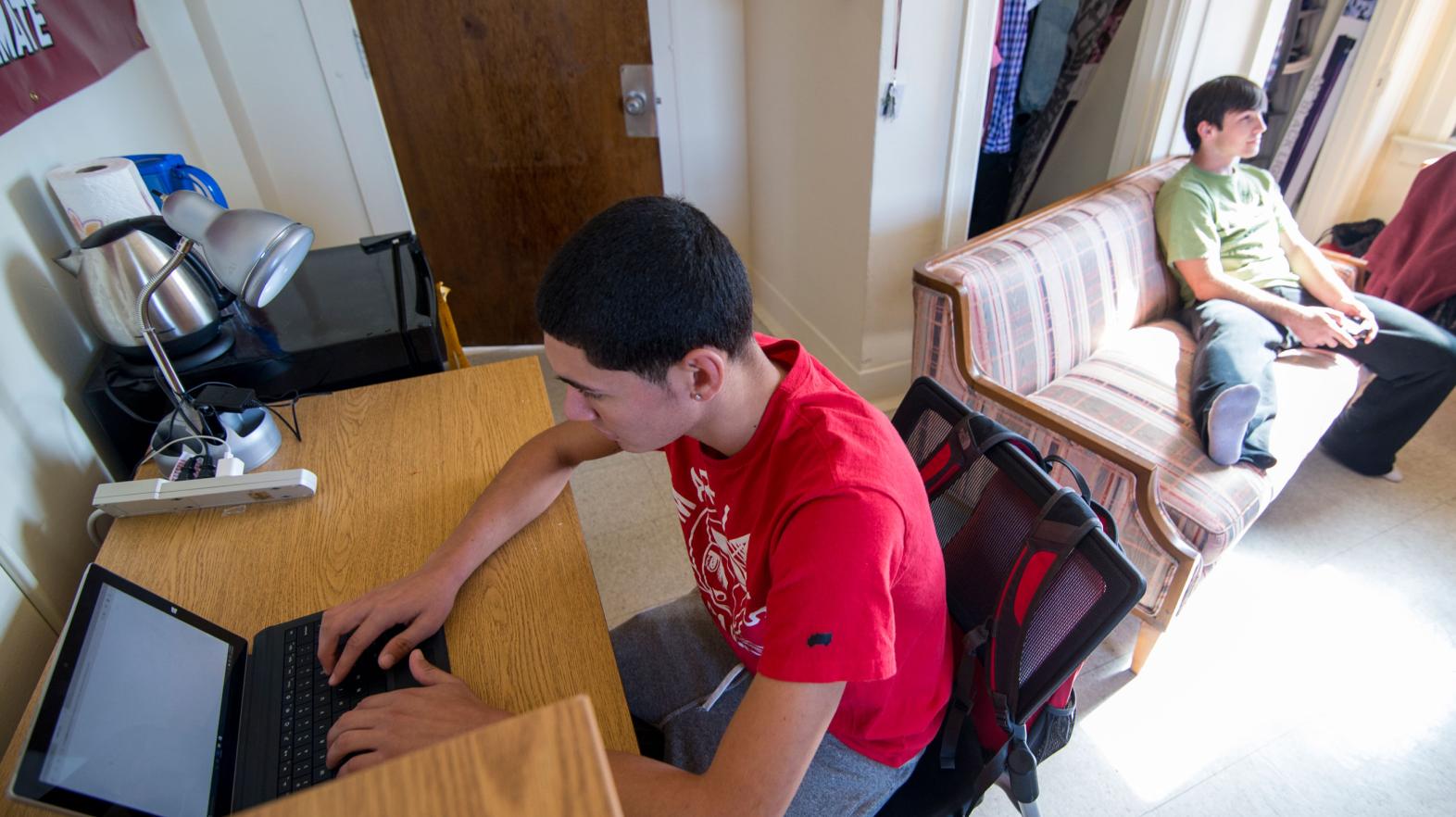 A student sites at his desk to work on his laptop while his roommate plays video games in their residence hall room.