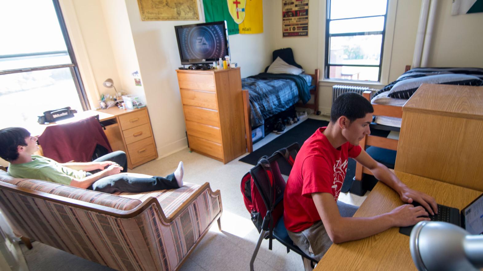 A student sites at his desk to work on his laptop while his roommate plays video games in their residence hall room.