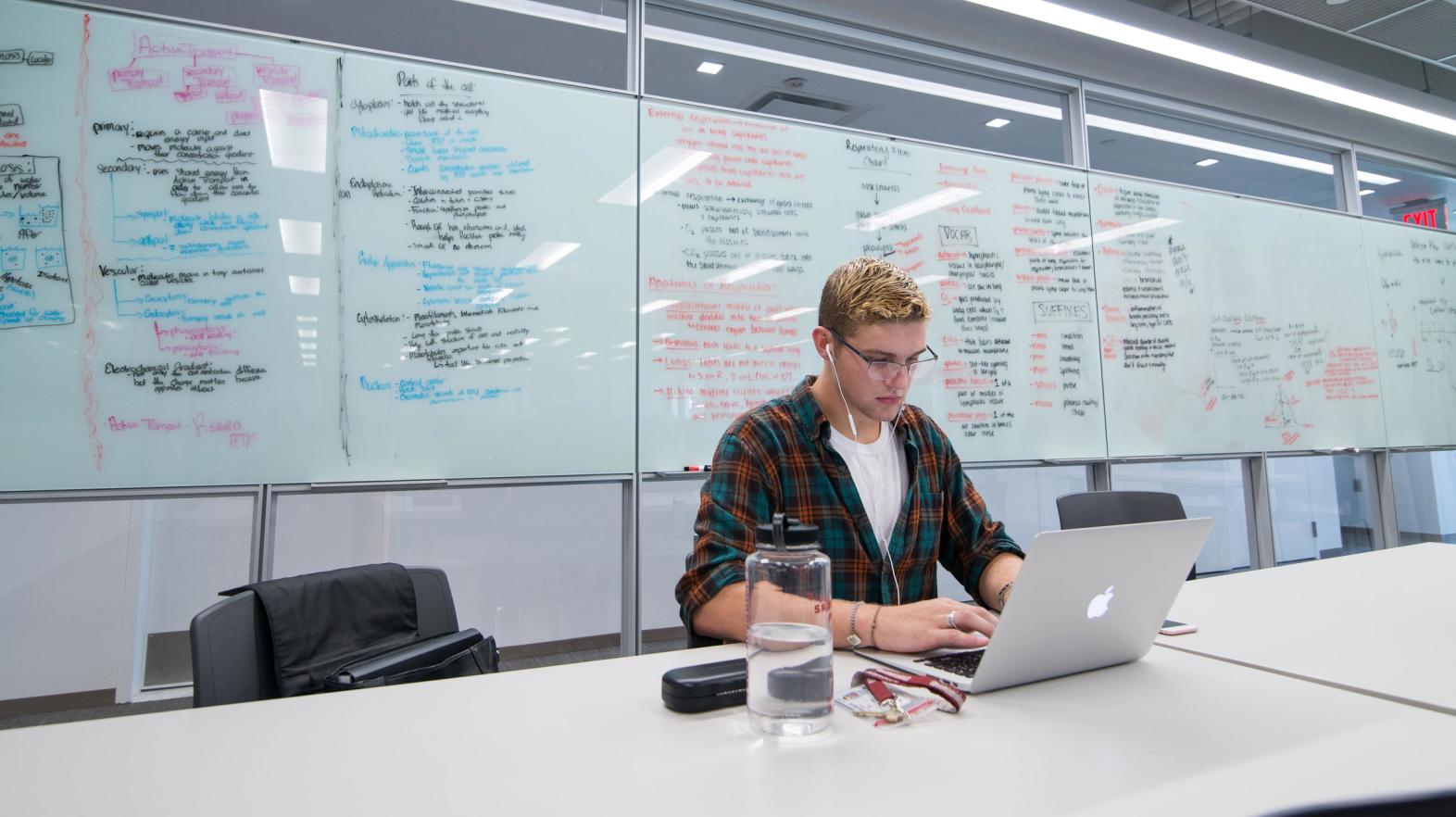 A male student types on his laptop as he sits in front of a white board with notes written on it.