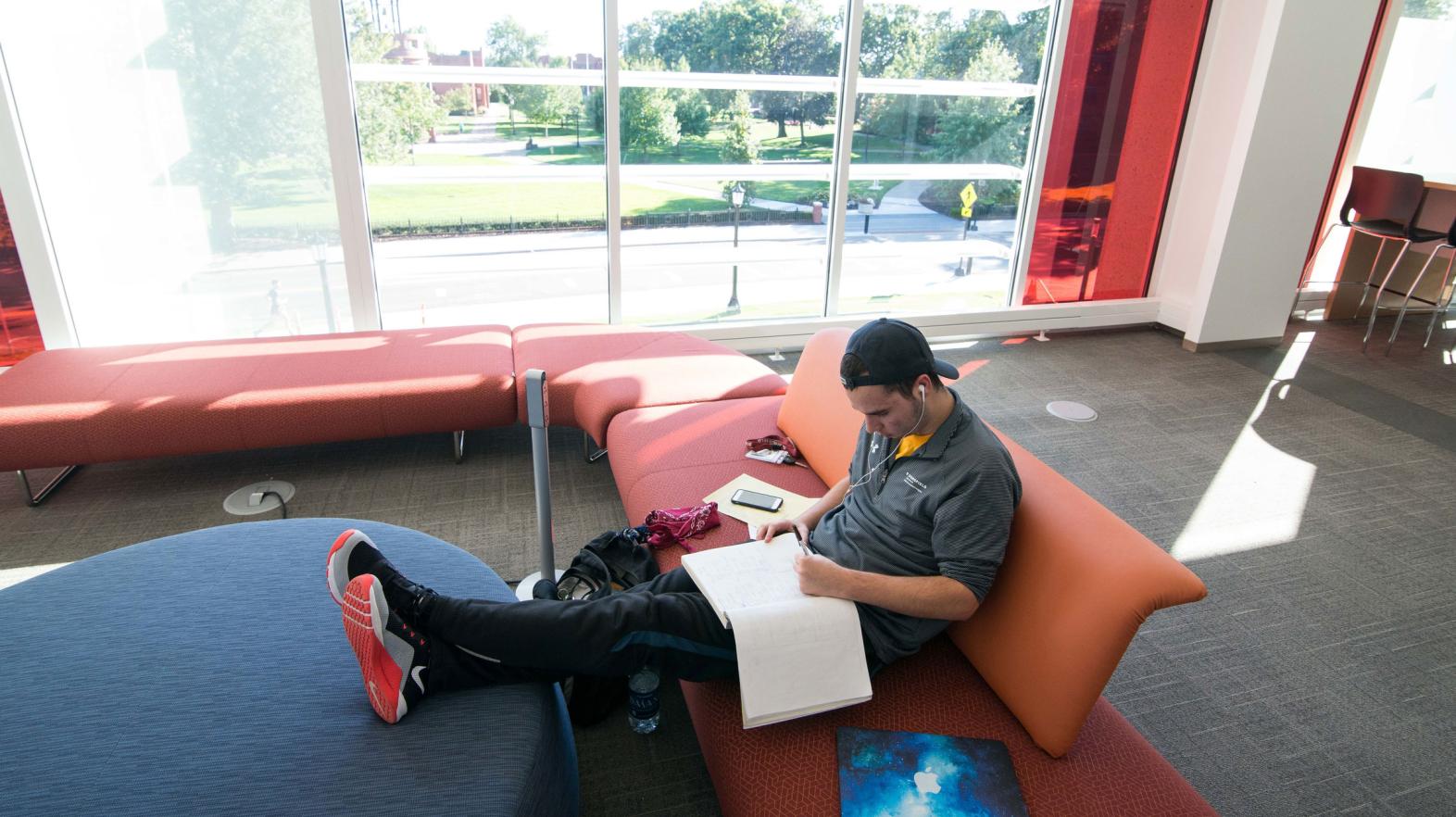 A male student reads from his notebook on a couch in the Learning Commons.
