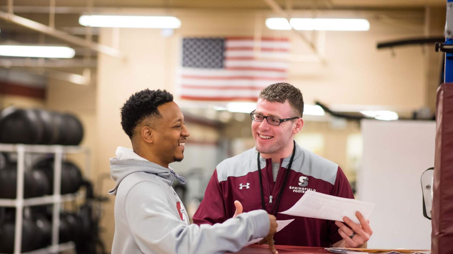 Two students talk and look over paper while in the weight room. 