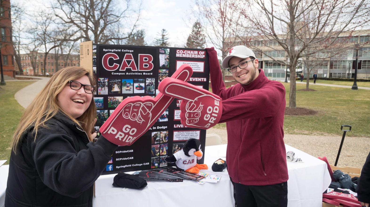 Two students stand with foam fingers in front of information about their club. 