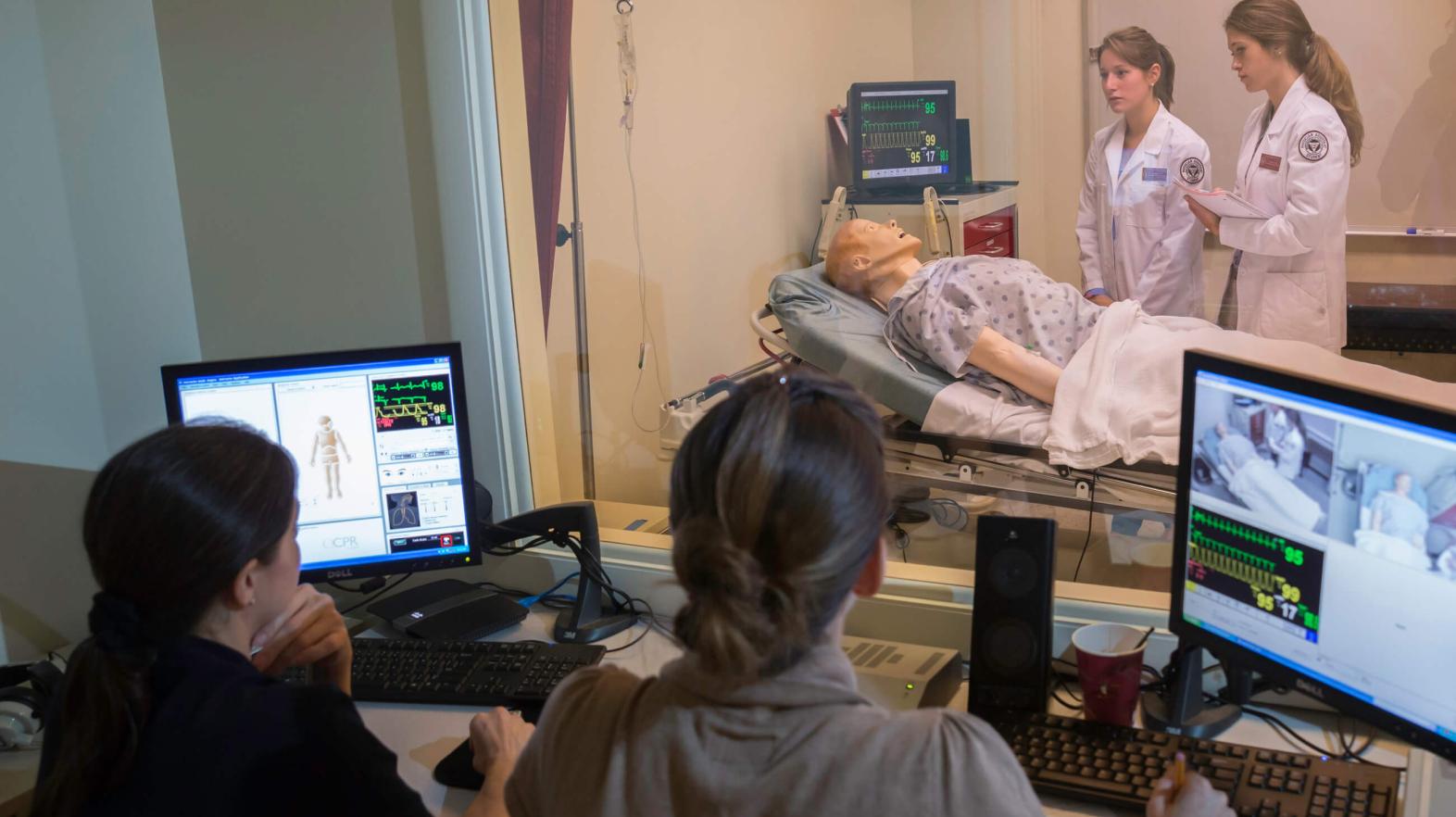 Two students observe two other students who are in the simulation lab.