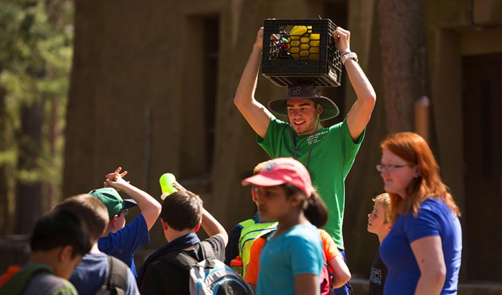 Springfield College student with a bucket of games and activities on their head surrounded by young children