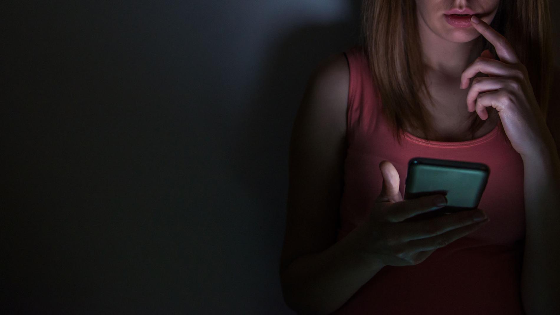 A girls sits in the dark, looking at her phone with concern. 