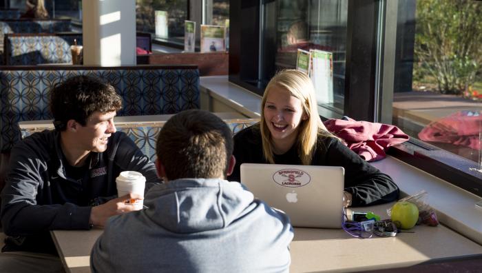 Students sit in the campus union and laugh while looking at a laptop