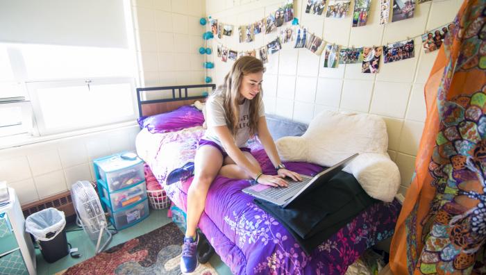 A young female Springfield College student sits on her bed in her residence hall while working on her laptop.